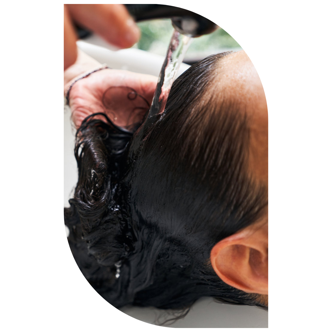 Person reclined with head in a wash basin as water runs through their wet hair while a hand rinses shampoo from the scalp.